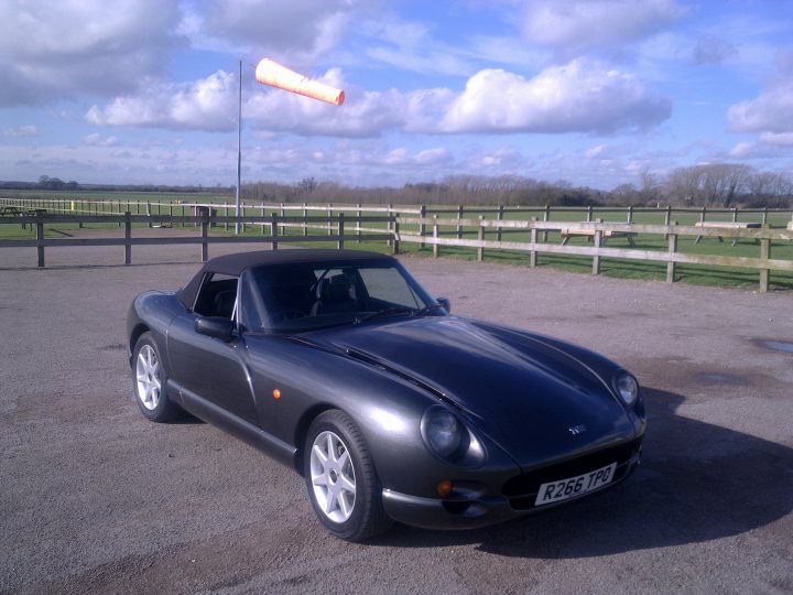 New Owner - Page 1 - Chimaera - PistonHeads - The image depicts an open landscape with a few clouds in the blue sky. The main subject is a small grey sports car parked on gravel. The car has a black soft top convertible roof which is down, showing the interior of the car. Behind the car, a wooden fence and some trees can be seen in the distance. Nearby, a tall flagpole with an orange flag is mounted on the gravel. There are no visible texts in the image. The scene appears to be quiet and peaceful, with no people or other vehicles in sight.