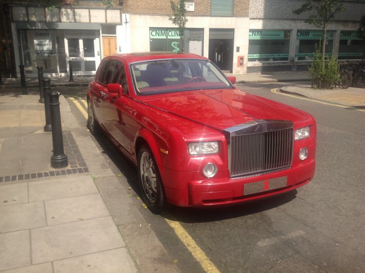 Spotted near my work I'm Fitzrovia... Amazing - Page 1 - Rolls Royce & Bentley - PistonHeads - The image portrays a vibrant city scene with a striking red Bentley car parked on the side of a street. The car, with its large grille and round headlights, is angled towards the camera, showcasing its luxurious design. Next to the car, a black lamp post stands tall, adding depth to the composition. The background consists of row houses, their storefronts reflecting a variety of shops, creating a lively urban tableau.