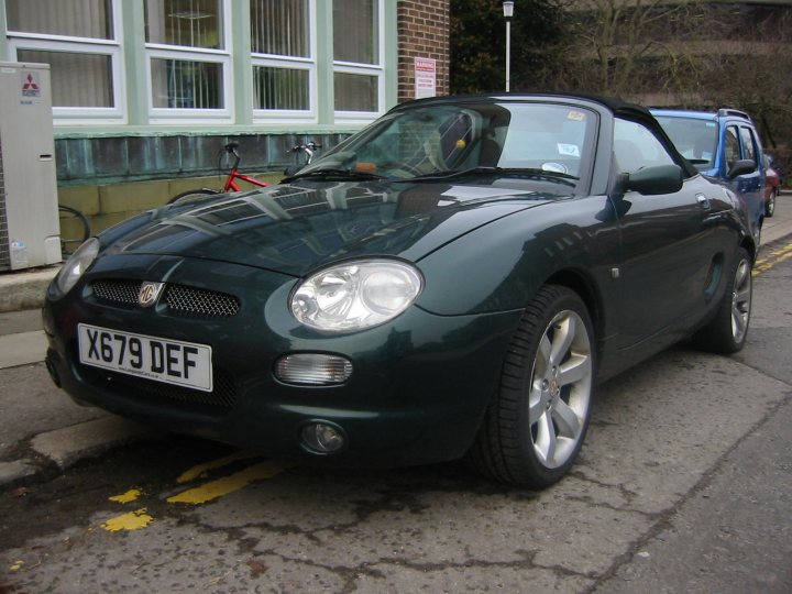 Pistonheads Mgf Exist Bargain - The image shows a green convertible parked on a street. It's a foggy day, and the car appears to be parked near the curb. A silver and blue car is visible in the background. The license plate of the green car reads "X679 DEF." On the left side of the image, a bicycle is parked against a wall. There is also a small sign near the car.