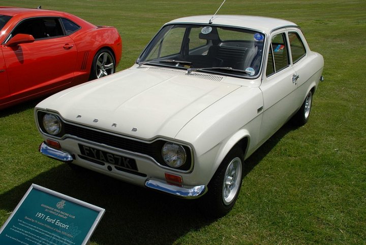 GRRC Lunch Club - Page 1 - Goodwood Events - PistonHeads - The image features a classic white car with a distinctive front grille positioned on a field of lush green grass. The car is a two-door saloon, equipped with a roll bar between the two doors, indicating possibly authentic or cop-themed alterations. Its design suggests it's from an older generation of vehicles. In the background, there's a glimpse of a red sports car, but it's less prominent compared to the white classic car. There's also a plaque displaying the words "1971 Ford Escort" inscription, further elaborating its identity as a '71 Ford Escort' among classic car enthusiasts.