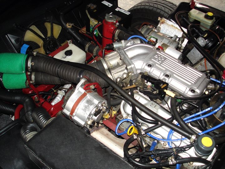 Identifying Pistonheads - This image shows a complex network of engine components, with a focus on various hoses and wires that are spread out across the frame. The engine's internal parts, such as the distributor cap and alternator belt, can be seen amidst the tangle of mechanical and electrical connections. In the background, part of a wheel or tire is visible, suggesting that the photo might have been taken under a car or a rack of tools. The organized chaos of the scene suggests a professional setting, perhaps a garage or a workshop.