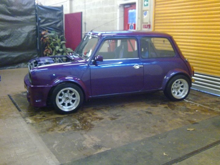 Pistonheads - The image features a vibrant purple small car, likely a mini cooper, parked in a warehouse or garage setting. The garage has a stone floor, and the car is oriented towards the left side of the image, with the rear of the vehicle facing the viewer. There are two distinct modifications visible on the car: a roll bar and a kame tail. The roll bar ensures safety, while the kame tail adds a playful and sporty touch to the vehicle's design. The car's positioning and the modifications suggest it might be used for performance driving or racing, given the small size and bright color, which are common in race or track-day vehicles. The garage itself provides a neutral background that accentuates the car's unique colors and details.