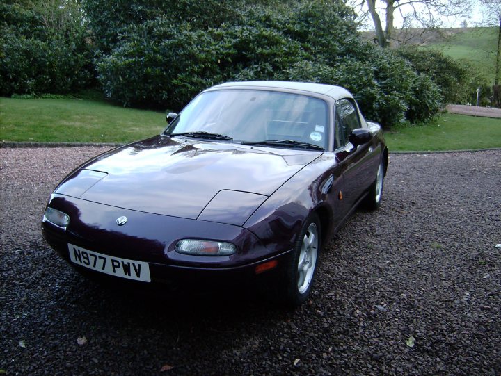 My Vr ltd comb A arrived today  - Page 1 - Mazda MX5/Eunos/Miata - PistonHeads - The image features an old, maroon-colored sports car parked on a gravel driveway. The vehicle has a convertible top and a chrome emblem on the front. In the background, there is a well-manicured lawn and a grassy embankment. The sky appears overcast.