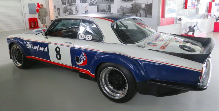 Pistonheads - The image features a vintage car, possibly from the 1970s or earlier, prominently displayed in a showroom setting. The vehicle is painted in a striking color combination of red and white with blue stripes on the hood. It bears a number "8" on the side and has a sponsor's name, "Kingland," written on it, suggesting its use in racing or motorsport events. There are also images of a person and a fire hydrant in the background, which appear to be related to the vehicle's context or history. The car is placed on a black surface, possibly a floor or platform, which contrasts with its vibrant colors.