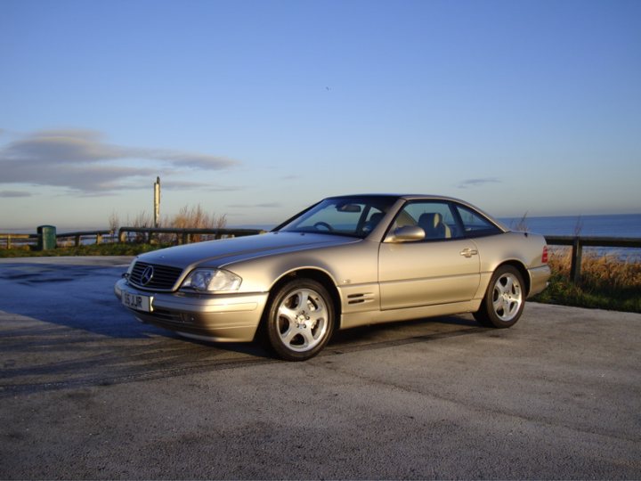 Show us your Mercedes! - Page 78 - Mercedes - PistonHeads - The image depicts a solitary, silver-colored car parked in what appears to be a clear, cloudless sky, suggesting early morning or late evening. The car is positioned on the right side of a highway, adjacent to a grassy area that is bordered by what looks like a sea beyond a fence. The vehicle's reflective surface mirrors the calm hues of the sky, enhancing the serene atmosphere of the scene.