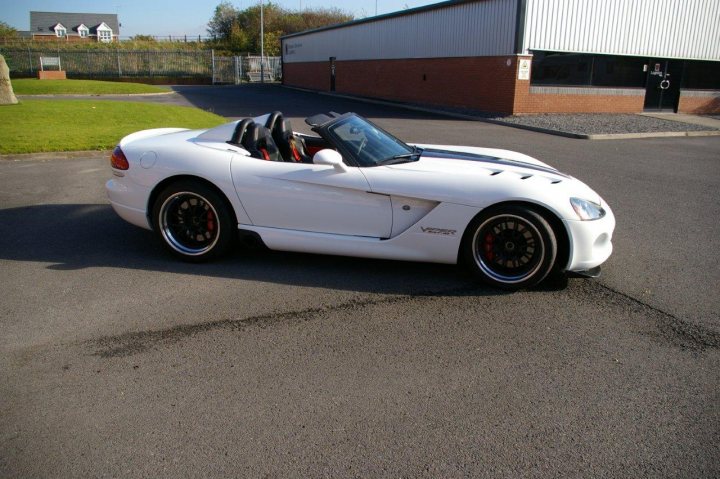 Rear Spoiler Or Not! - Page 1 - Vipers - PistonHeads - The image shows a white convertible sports car parked on an asphalt surface. The car's top is down, revealing two black seats with pink piping. The vehicle has red rims and a side exhaust pipe. The car is positioned in the foreground with a building whose windows reflect a faint blue sky. The ground appears wet in places, suggesting recent rainfall.