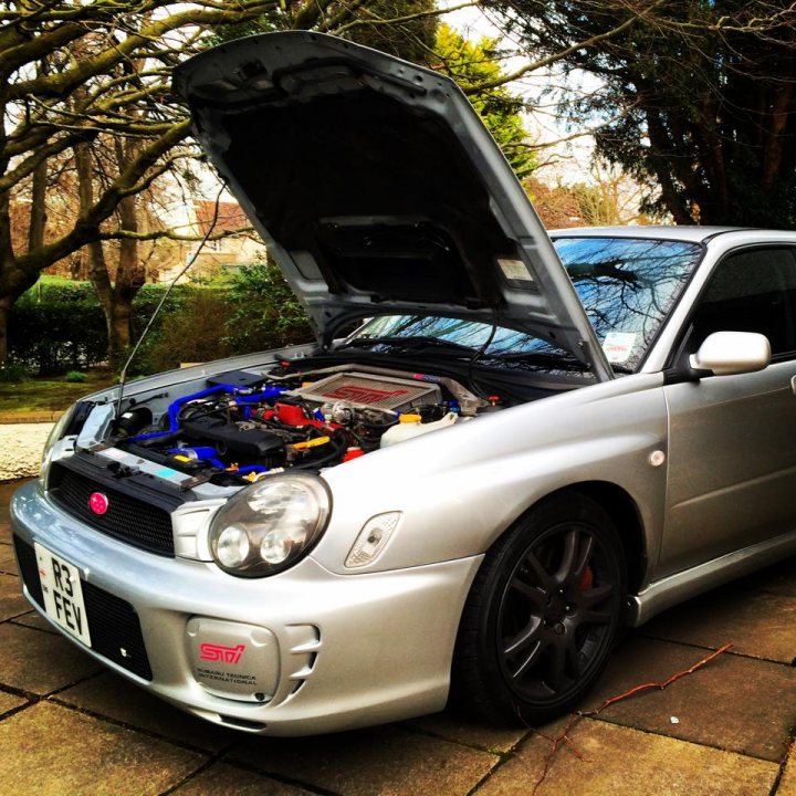 Bugeye JDM Sti - Page 1 - Subaru - PistonHeads - This image captures a scene in a driveway where a silver-colored car with a modified suspension system and a lifted hood is prominently displayed. The car's engine is visible, revealing its blue and black components. It's parked on a paved surface which contrasts with the natural surroundings, including what appears to be a forested area in the background. The day is overcast but the hood of the car has been opened, possibly for inspection or modification purposes. The overall ambiance gives off a sense of luxury and mechanical interest.