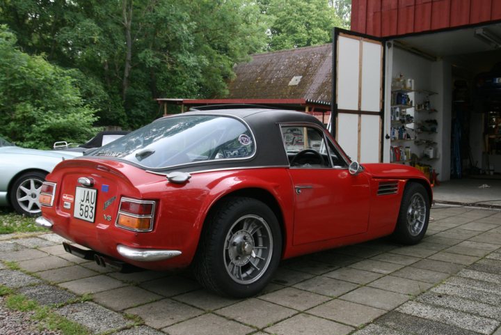 Early TVR Pictures - Page 36 - Classics - PistonHeads - The image shows an old red vehicle with a convertible top parked on a driveway. The vehicle is parked in front of a white garage with a red roof, and there is a green car parked in an adjacent spot. The driveway is lined with a grass verge and the setting appears to be suburban or rural, as there are trees visible in the background. The style of the image is a straightforward, non-artistic photograph capturing the scene.