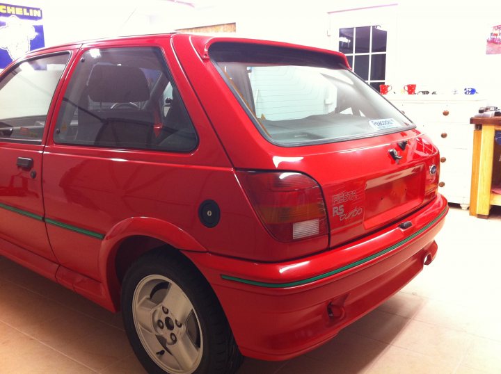 Fiesta RS Turbo Restoration - Page 13 - Ford - PistonHeads - The image shows a red car with a black rear notice, parked indoors with a white wall in the background. There are several cups and plates on cabinets visible in the background, suggesting the setting might be a kitchen or a dining area. The car appears to be a hatchback, and its reflective surface gleams under the indoor lighting. A small sticker is placed on the rear window of the car, adding a personal touch to its otherwise shiny exterior. The car is positioned facing the camera, allowing a clear view of its profile.