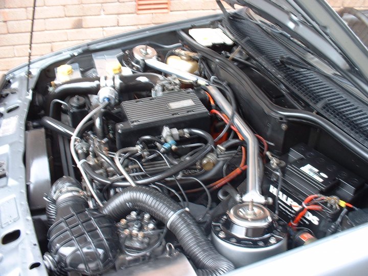 Turbo Technics Sierra Pistonheads Xrx - The image shows a close-up view of the engine compartment of a car, revealing several components. The engine is densely packed, with various wires and parts visible. Notably, there's a large black battery with red wiring connected to it, which appears to be the primary source of power for the vehicle. There are other components including what appears to be a metal box with 'Tick' written on it and a smaller black battery nearby. The internal view of the engine reveals an array of tubes, wires, connectors, and gaskets that are part of a complex mechanical system. The metal could be a firewall or part of a protective cover. The complexity of the machinery highlights the sophistication of modern vehicle design.