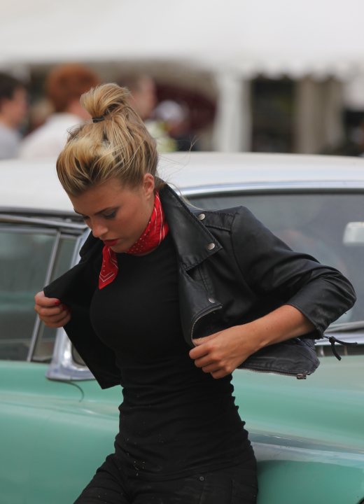 Pistonheads Hunnies Goodwood - The image captures a moment of a woman adjusting her vibrant red bandana worn against her dark hair. She is standing next to an equally striking green vintage car. The focus is on her attire and action, suggesting she might be preparing for a drive. In the background, blurred figures hint at the presence of several people, but the main scene is centered around the woman and the car. The image exudes a vintage aesthetic, likely evoking a nostalgic feeling.