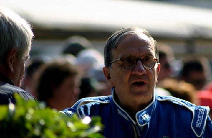 Post your Pics, Celebrities at  FOS 2011 - Page 1 - Goodwood Events - PistonHeads - The image is a close-up photograph of an older individual in the foreground wearing glasses and a blue garment with a white stripe. The person appears to be outdoors with others around, though their face is in focus, making it the main subject. There seems to be a car and foliage in the background, suggesting a casual, possibly outdoor event. The lighting suggests it might be morning or late afternoon, given the direction of the shadows. The overall style of the image is realistic, capturing a moment with some candid emotion, as indicated by the person's open mouth.