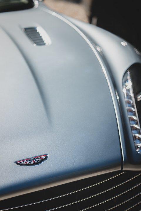 How about an Aston photo thread! - Page 173 - Aston Martin - PistonHeads - The image shows the hood of a silver or light gray car. The vehicle has a slightly curved hood that reflects a bit of steel blue or black at its edges. On the left side of the hood, there's an emblem with a logo that includes a clock face with stripes emanating from the center of the circle. The design is sleek and modern, suggesting a sports car or a high-end vehicle. The photograph is taken at a sharp angle, emphasizing the shape and design of the car's hood.