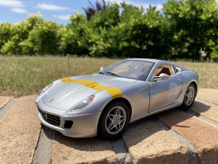 A car with a surfboard strapped to the back - The image showcases a silver sports car toy with yellow and black stripes on its side, placed against the backdrop of a brick wall. The car is positioned slightly to one side, with a clear view of its sleek design and shiny exterior. It's situated in an outdoor setting, under what appears to be a sunny sky, as indicated by the brightness and shadows cast across the scene. A hint of greenery can be seen in the top left corner, adding a touch of nature to the overall composition.