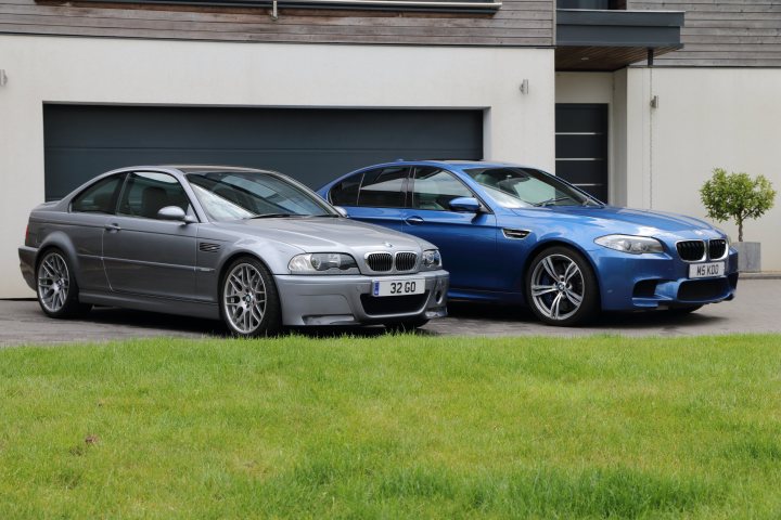 M3 CSL and M5 - Page 6 - Readers' Cars - PistonHeads UK - The image is a photograph of two sports cars parked side by side on a driveway. The car on the left has a silver metallic finish and appears to be an older model, possibly a BMW. Its counterpart on the right is in a vibrant shade of blue, also resembling a BMW, and has a more modern design. Both cars are parked facing the viewer, and there is a house with a visible garage door in the background. The setting suggests a residential area, with the driveway being made of concrete or asphalt.