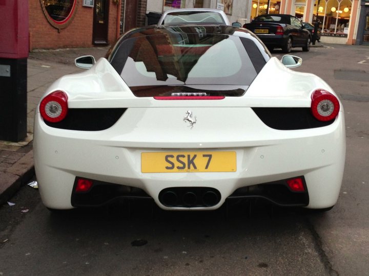 Spotted 2013 - Page 1 - Northamptonshire - PistonHeads - The image features a white Ferrari with a visible red taillight, driving on a street lined with parked cars. The car is positioned behind a pole on the street. In the background, there is a brick building with a window that reflects the scenery. The Ferrari's license plate reads "SK7," suggesting that this is a personal vehicle as opposed to a promotional or company car. The overall scene conveys a sense of urban environments and high-end automobiles.