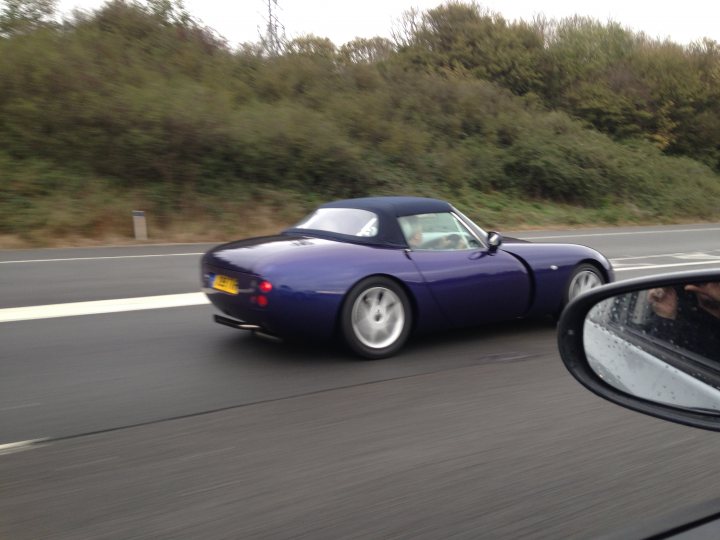 A car is parked on the side of the road - Pistonheads - The image depicts a striking, deep purple convertible sports car gliding down a real-life highway. The car, with its sleek design and vibrant color, stands out against the solid lines of the road. In the driver's seat, you can see the profile of the driver's head. The environment shifts between the car and the natural landscape, suggesting movement along the highway. The reflection of the car's back and the foliage in the mirrors adds a unique perspective on the journey.