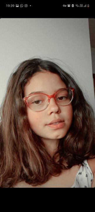 A woman in a red shirt and a red tie - The image is a screenshot from a video call or messaging app. It shows the profile picture of a person, who appears to be a woman with dark hair and glasses. She is looking directly at the camera with a neutral expression. The background is plain and does not offer any context or setting. The lighting in the photo is soft and evenly distributed, suggesting an indoor environment.