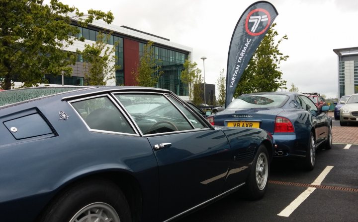 A car parked next to a parking meter - Pistonheads - The image shows a parking lot during what appears to be a cloudy or overcast day. There are two luxury cars parked side by side: a blue convertible on the foreground and a blue coupe on the background. Between these cars, a flagpole stands with a banner attached, although the text on the banner is not clear. The cars have aftermarket modifications, indicated by the presence of spoilers, which give the vehicles a sportier look. There are no visible texts or branding that confer specific information about the cars or the event.