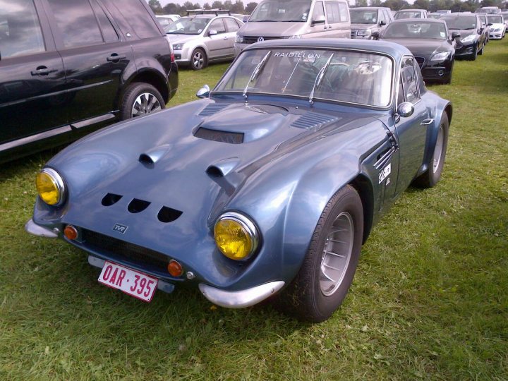 Early TVR Pictures - Page 14 - Classics - PistonHeads - This image depicts a vintage racing car styled like a rally car, parked on grass among other cars and with people in the vicinity. It has a textured blue body with yellow headlights and a license plate reading "OAR 395". The car's design features two prominent front air intakes and a sloping rear deck spoiler. The surrounding grass suggests an outdoor setting, possibly a European event or show.