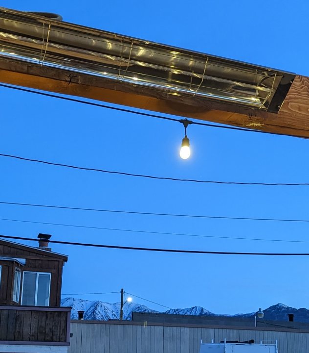 Pistonheads - The image captures a serene night scene in what appears to be a mountainous area. Dominating the frame is a rustic, wooden building with an overhead roof. A metal drainage pipe runs across the center of the roof, adding an industrial touch to the otherwise traditional structure. 

A string of lights hangs from this pipe, casting a warm glow over the scene below. The building seems to be part of a larger complex, as evidenced by a street light standing prominently in the foreground on the left side of the image. 

The background features a vast expanse of dark sky, punctuated by distant mountain silhouettes under the stars. On the right side, there's a telephone pole standing tall against the night sky. The entire scene is bathed in a cool blue hue, lending an air of tranquility to this idyllic setting.