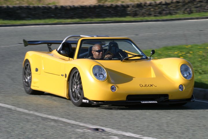 Kit Pistonheads Supercar - The image shows a scene of a black and yellow Porsche sports car driving on a rural road. The car is at an angle to the camera, indicating motion. The car's sleek design and vibrant colors stand out against the backdrop of the winding road and the greenery on either side. The driver, wearing sunglasses, is visible and appears focused on the road ahead. The overall atmosphere of the photograph suggests a day of joyful driving on a beautiful country road.