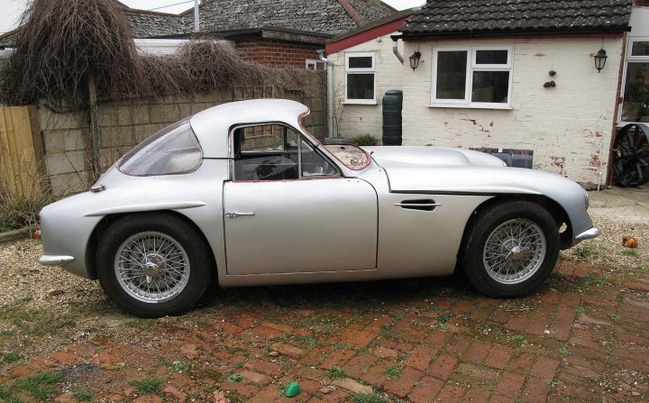 Early TVR Pictures - Page 75 - Classics - PistonHeads - This image captures a vintage sports car in a charming outdoor setting. The car, which appears to be a silver convertible with a white soft top, is the main focus of the scene. It is parked in a driveway in front of a delightful old-style cottage, complete with cream walls, a terracotta roof, and a cozy-looking chimney. Surrounding the car and the cottage is a neatly trimmed lawn, adding to the idyllic atmosphere. A wall on the side of the vehicle displays hints of greenery, suggesting a lush garden or yard. The composition of the photo suggests a peaceful day spent in a well-kept, nostalgic environment.