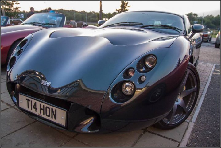 TVRCC + Guests Berkshire June Meet - Page 1 - Thames Valley & Surrey - PistonHeads - The image shows a close-up of a high-end sports car, with only the front section of the vehicle visible. The car is a silver, two-door convertible, and it features a sleek design with a prominent front grille and large round headlights. The vehicle is parked on the side of a road, with a blurred background indicating it might be a windy day. The license plate of the car reads "TI4 HON."