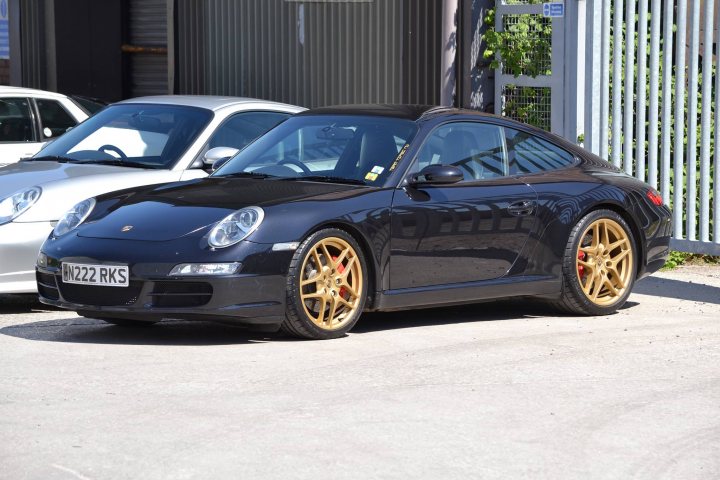 lowering a 997 - Page 2 - Porsche General - PistonHeads - The image shows a black Porsche Cayenne parked outdoors. The car is positioned at an angle, facing slightly to the right. It features gold wheels and a visible license plate. In the background, there are other cars parked in what appears to be a parking lot or garage. The ground is covered with concrete, and the sky is overcast. The setting suggests this photo was taken during the day under cloudy weather conditions.