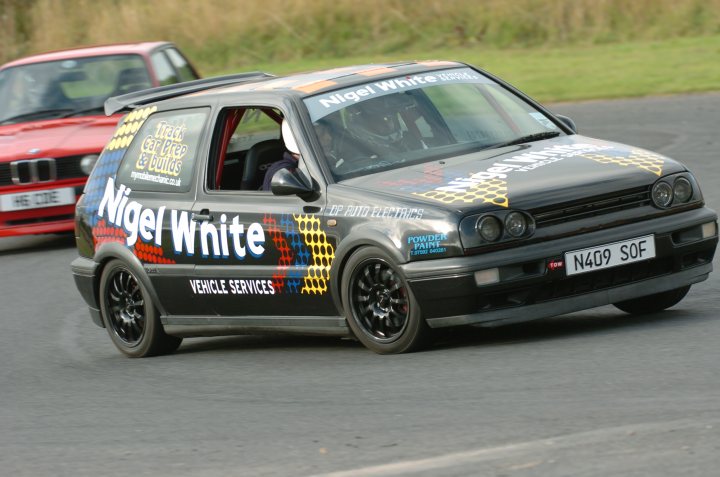 Teesside Autodrome Next Week (25th Sept.) - Page 1 - North East - PistonHeads - The image captures an intense moment on a race track. A gray, silver, and black car speckled with a "Nigel White" advertisement is seen drifting, a testament to the skill and control of the driver. In the background, a red car follows relentlessly, adding to the dynamic of the scene. The vehicles are on a track lined with green grass, under a bright blue sky. The blur of motion and the focus on the foreground car give a sense of speed and excitement.