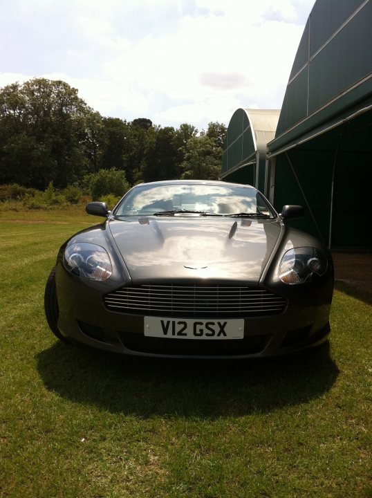 Pistonheads - In the image, there's a sleek, black Aston Martin car parked on a grassy field. The car is oriented towards the left side of the photo and bears a license plate that reads "V12 GSX". The car's metallic sheen contrasts with the natural hues of the green field and the overcast sky above, which adds a dramatic effect to the scene. In the background, there's a large white building with a green roof, providing a backdrop for this luxurious vehicle. The car's position on the grassy expanse gives the impression of it being in an open, spacious area.