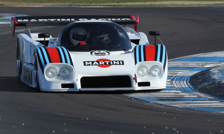 Racing Historic Donington Pistonheads - The image features a racing scene with a unique vehicle known as the Martini Racing machine. The vehicle is mostly white with blue and red stripes, indicative of the Martini branding. It appears to be a heavy-duty racing car, possibly used in endurance races, with features such as a large front bumper and the number six on the side. In the cockpit, a driver is visible, donned in a protective racing suit, wearing a helmet with a white visor. The racing car is in action, driving on a race track with a clear cast-off surface.