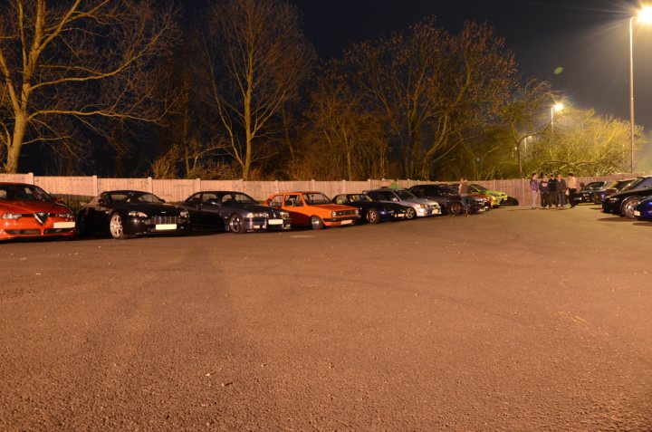 Essex March Pistonheads - The image shows a parking lot filled with several cars, both in motion and parked. The cars vary in color and model, creating a diverse scene. The lot is bordered by a wooden fence, and the night sky above suggests early evening or night time. At the far end of the lot, there is a line of trees, providing a natural backdrop to the park area. Lighting floods the lot from the right side, illuminating the area and emphasizing the silhouettes of the automobiles.