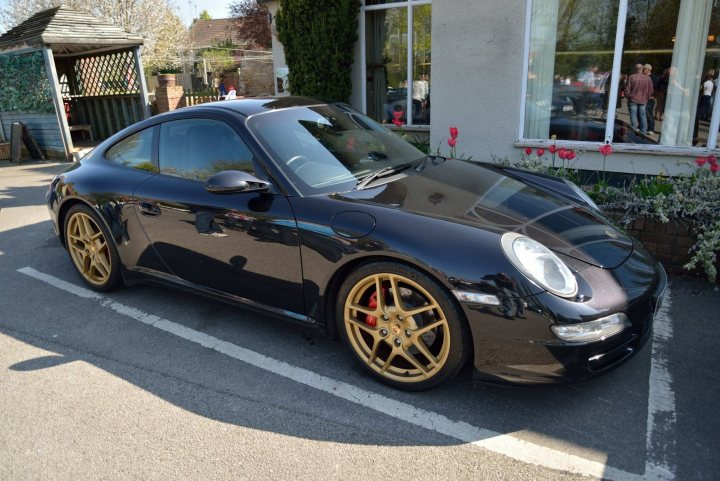 997.1 C2S Wheel colour - Page 1 - 911/Carrera GT - PistonHeads - The image shows a stylish black Porsche parked on the side of the road. The vehicle features gold wheels that add a touch of luxury to its appearance. It is parked outside a building that has large windows, and there is a red flower planter adorned with red flowers nearby. To the left of the vehicle, there is a structure made of wood and glass. The scene suggests it might be a pleasant day, as trees can be seen in the background and the shadows on the ground indicate that the sun is out.