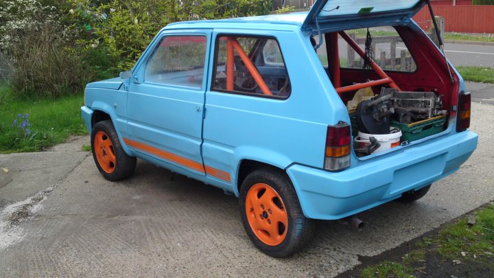 Sublime Pandamonium II - Page 23 - Alfa Romeo, Fiat & Lancia - PistonHeads - The image showcases a small blue car parked on a sloping driveway. The car has an open hatchback, revealing a variety of items stowed within. Notably, there appears to be a lawnmower and a plastic container, suggesting that the car owner might be preparing for some outdoor work or cleaning. The surrounding area is tranquil, with a garden and a tree visible, adding a touch of green to the scene. The driveway on which the car is parked appears to be made of concrete, and a sidewalk can be seen in the background.