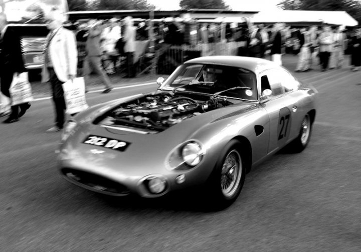 Revival - First Time and Excited - Page 1 - Goodwood Events - PistonHeads - The image is a sepia-toned photograph of a classic car, specifically a vintage sports car, positioned on a street for display. The car appears to be in good condition, with various parts and panels missing or exposed, revealing a complex engine and mechanical components. Surrounding the vehicle, there's a blurred crowd of people who seem to be milling around and observing the car, suggesting this may be a public event showcasing vehicles. The setting seems outdoors, and there are buildings that provide a sense of an urban environment. The photograph has an artistic touch with the selective color treatment, focusing on the car while rendering the rest in black and white.