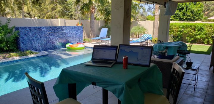 Share your HOME WORKING workstation environment - pics - Page 19 - Computers, Gadgets & Stuff - PistonHeads - This is a photograph of an outdoor dining area adjacent to a swimming pool. The image captures the scene from the perspective of someone inside the house, looking out through a sliding glass door onto the patio. On the left side of the photo, there are tables covered with blue tablecloths and chairs, suggesting a recent or upcoming gathering or event. There's also some electronic equipment on one of the tables, possibly related to work or a presentation. The pool area is furnished with lounge chairs under an umbrella, inviting relaxation in the sun. The backyard features a blue tiled wall and palm trees, contributing to a tropical atmosphere. There are no people visible in the image.