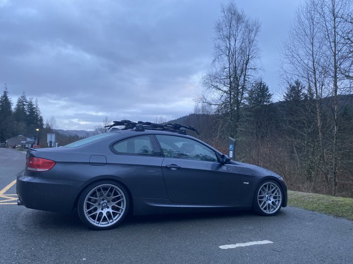 My brave pill; E92 BMW 335i with the infamous N54 engine - Page 43 - Readers' Cars - PistonHeads UK - The image shows a dark blue BMW car parked on the side of a street, which appears to be a country road. The car is a convertible model with a hardtop, and it features a black roof rack. The vehicle has custom rims and a visible sunroof. It's currently raining, as indicated by the overcast sky and wet appearance of the car. There are no people visible in the image. The surrounding area appears to be rural or semi-rural, with trees and a hint of a body of water on one side.