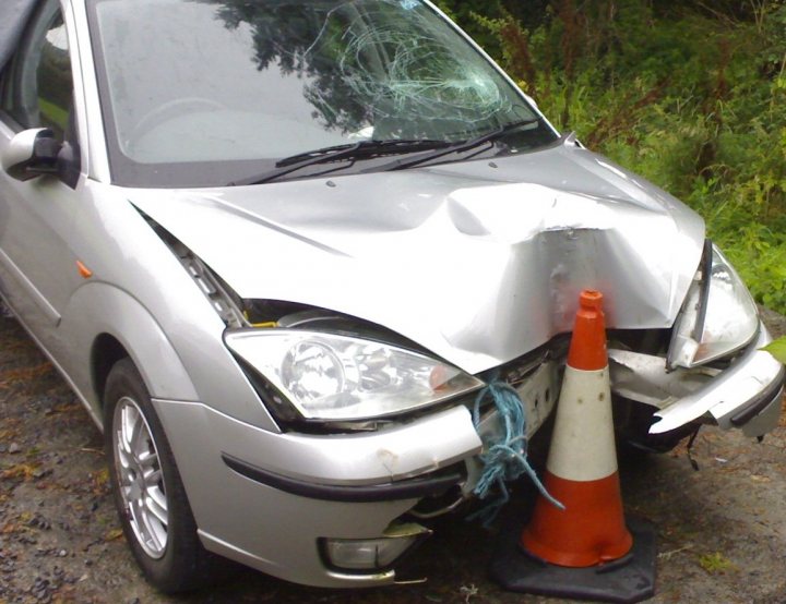 Show us your crash pics!! - Page 27 - General Gassing - PistonHeads - This image captures the aftermath of a car accident. An aluminum hatchback is in the foreground, with a white and orange road cone wedged into its damaged front bumper. The car has sustained significant front-end damage, including a smashed hood and front grille. The surrounding scene is somewhat blurred but shows a grassy area with patches of shrubbery, suggesting the aftermath of the accident. The presence of debris and the state of the car suggest a severe collision.