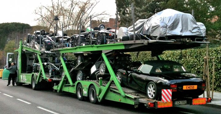 Car Transport Spain to UK and back - Page 1 - Spain - PistonHeads - The image shows a large, flatbed truck carrying a diverse collection of vehicles. Atop the truck rests a sports car, followed by various motorcycles, creating a colorful assortment of vehicles. The trailer beneath the truck bed appears to be equipped with a green frame, possibly for transporting fragile or low-profile cars. The setting of the image consists of a quiet street lined with store fronts, suggesting a commercial or industrial area. A lone individual can be spotted at the far left of the image, walking on the sidewalk.