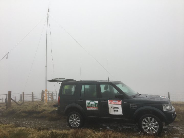 The 2017 Rallying thread - Page 32 - General Motorsport - PistonHeads - This image shows a car positioned on a road with a backdrop of a foggy landscape, possibly on a hill or a mountainous area. The car appears to be a black SUV and has a sign mounted on the hatch, with the text "WALES ELECTRIC SPORTS TEAM" visible. The fog is dense, reducing the visibility of the surroundings, but there are indications of what could be a metallic fence or some other form of barrier in the background on the left side of the image. The sky is overcast, reinforcing the sense of a misty or damp setting. The car's location and the surrounding environment suggest that it could be raining or about to rain.