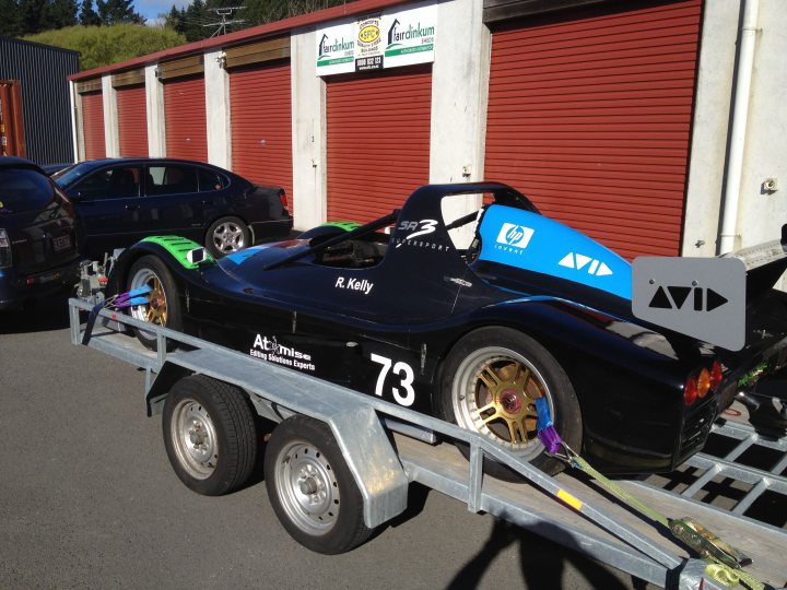 A motorcycle is parked in a parking lot - Pistonheads - The image captures a scene in a parking lot where a car carrier trailer is transporting a racing car that is predominantly black with white accents. The trailer appears to be sturdy and built for transporting sports cars, while the car, labeled with the number 73, is in front of a red building with a green sign. This suggests that it might be a race car preparing for a competition or exhibition. Another car, a blue sedan, can be seen parked in the background, adding to the atmosphere of a busy parking lot.