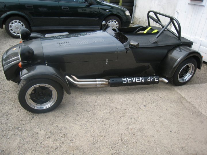 A motorcycle parked in a parking lot next to a car - Pistonheads - The image features a distinctive vehicle with the name "SEVEN JEREMY" prominently displayed on its side, suggesting it is a personalized car or tractor. The vehicle sits on a gravel driveway, with a muddy area around it, indicating recent outdoor activity. This unique vehicle appears to be based on a car with a prominent front end and large rims, but instead of the usual car body, it has an enclosed cockpit, reminiscent of a one-person car or land yacht. In the background, another car is partially visible, adding contrast to the main subject. The image exudes a sense of antiquity and individuality.