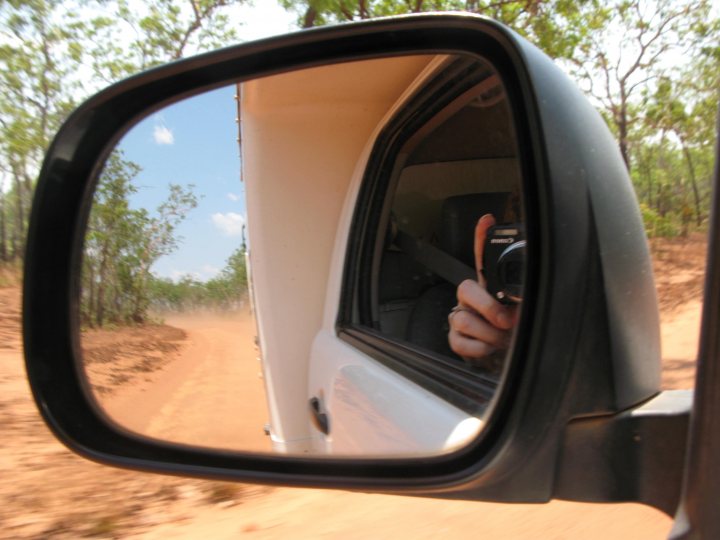 A reflection of a dog in the side mirror of a car - Pistonheads - This image captures a moment while driving a white vehicle down a dirt road. The viewer can see through the rearview mirror of the vehicle, where a person's hand is holding a cell phone to take a photo. The person is captured from behind, with the natural landscape stretching out down the dusty dirt road ahead. The surrounding environment includes trees and a calm atmosphere.