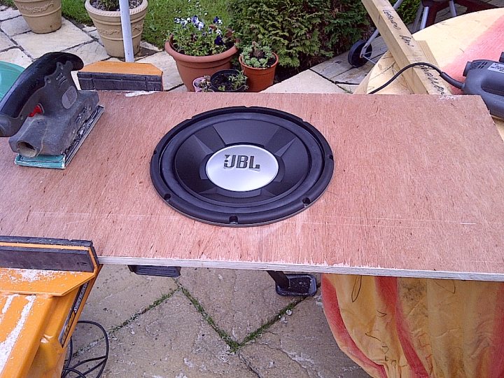 Music in a field - Page 1 - Tents, Caravans & Motorhomes - PistonHeads - In the center of the image, a black JBL speaker is mounted into a wooden rectangular plank. The speaker is only connected by one visible screw to the rest of the work area. There is some yellow wood or exterior grade paint on a nearby table. To the left, there is a metal vice clamp, indicative of a workspace. Background elements include various potted plants and extension cords, suggesting outdoor or accessory use.