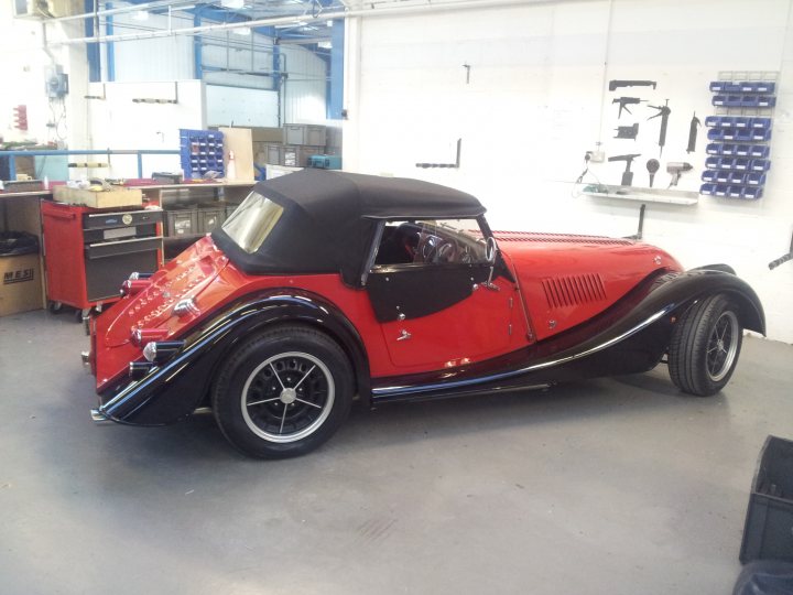 A couple of photos from a recent Factory tour... - Page 1 - Morgan - PistonHeads - This image shows a well-maintained, vintage-style car in a workshop setting. The car has a striking red and black paint scheme with gold accents. Its bodywork is smooth, and there are additional wheels visible at the back, which are likely spare tires. The interior features a black soft top, adding to the classic aesthetic. The vehicle is parked on a concrete floor, and a variety of tools and equipment are evident in the background, indicating that this might be a professional car repair or restoration shop.