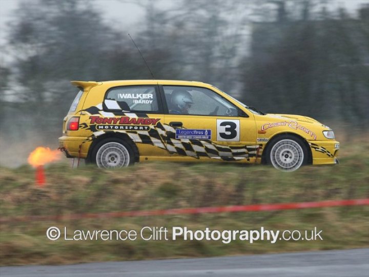 Pistonheads - This is a dynamic action shot, showing a yellow race car with a vibrant checkered pattern on its sides, predominantly black and white, racing down a wet track. The vehicle has the number 3 prominently displayed on its side and a name, "Walker," also visible. It is captured in motion, with smoke emanating from the back, indicating high speed. The car is equipped with a large rear wing, which is a standard feature for improved aerodynamics in racing. The image is credited to Lawrence Clift Photography, suggesting that the photographer is a professional.