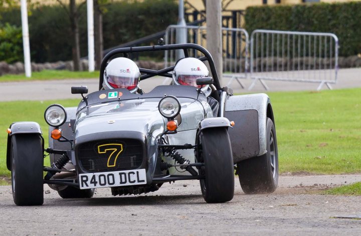 Pistonheads - In the image, two individuals are seen riding a silver sidecar bus, specifically designated as a Mobil Twinning Car No. 6400 and displaying the number 7. The sidecar bus is driving on a road. In the background, there's a chain-link fence to the right side of the frame. Both riders are wearing helmets, and the bus appears to be a convertible type with a distinctive silver finish.