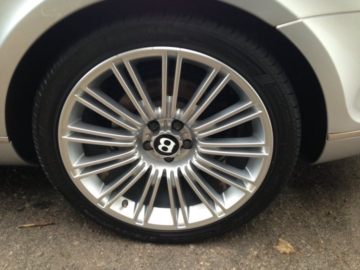 I’ve put my football boots on & bought a Bentley GT Speed - Page 1 - Rolls Royce & Bentley - PistonHeads - The image showcases a close-up view of a car's wheel, which is prominently displayed against a neutral backdrop. The wheel is a five-spoke silver alloy, with each spoke connected to a hub adorned by a large, black emblem. The tire itself has a noticeable tread, and the overall condition of the wheel and the tire's tread appears to be in good shape. The background, though blurred, indicates that the vehicle is parked on a concrete surface.