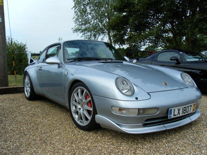 Breakfast Meeting, The Hare, Roxwell 4th June - Page 1 - Kent & Essex - PistonHeads - The image features a silver Porsche sports car parked on a gravel surface. The car has a sleek design with distinctive round headlights and silver bumpers. It's positioned with its hood turned slightly towards the viewer. In the background, the sky is partly cloudy, and there are other vehicles present, one with a contrasting black roof. The setting appears to be an outdoor parking area, possibly a car enthusiast's driveway.